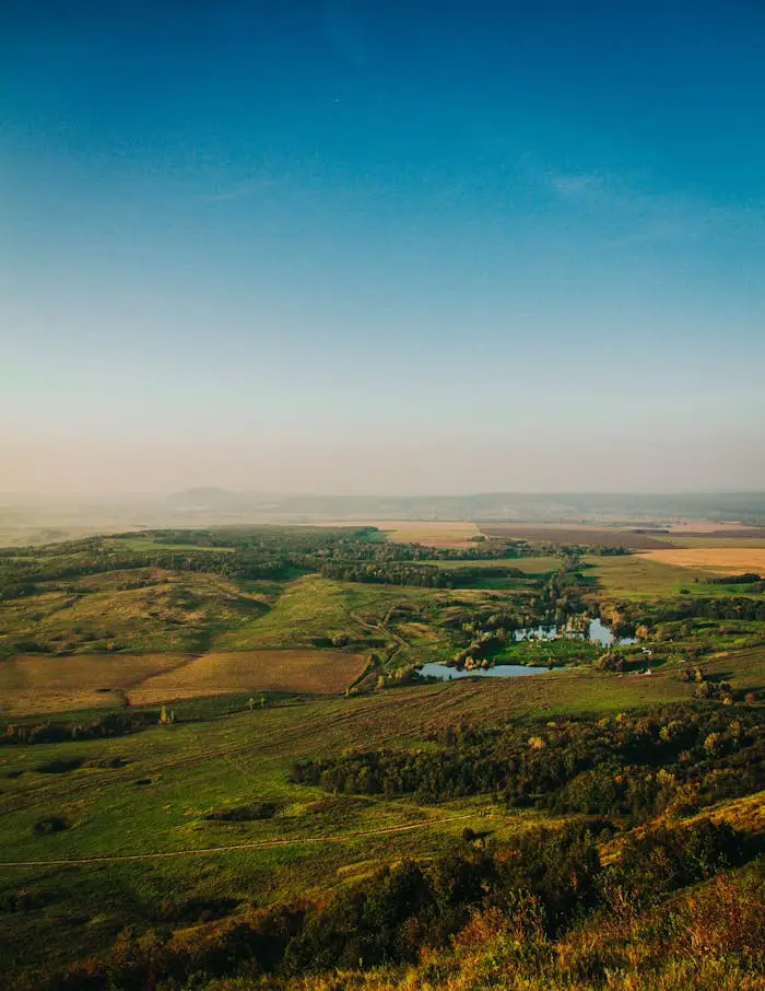 An expansive aerial view of the countryside with rolling green hills, golden farmland, and a serene pond, all under a vast, clear blue sky during the golden hour.